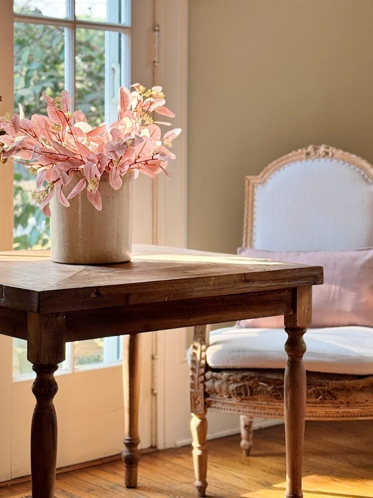 A wooden table with a pot of pink flowers next to an upholstered chair in a sunlit room.