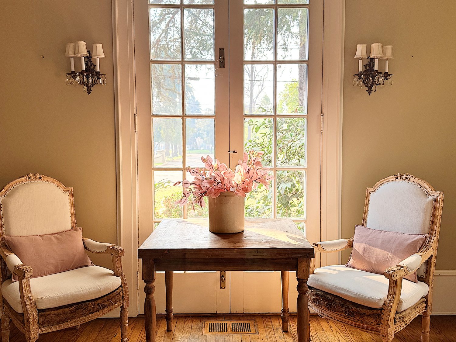 A cozy sitting area with two upholstered armchairs, a wooden table, and a vase of pink flowers, set against large glass doors leading to a garden view.