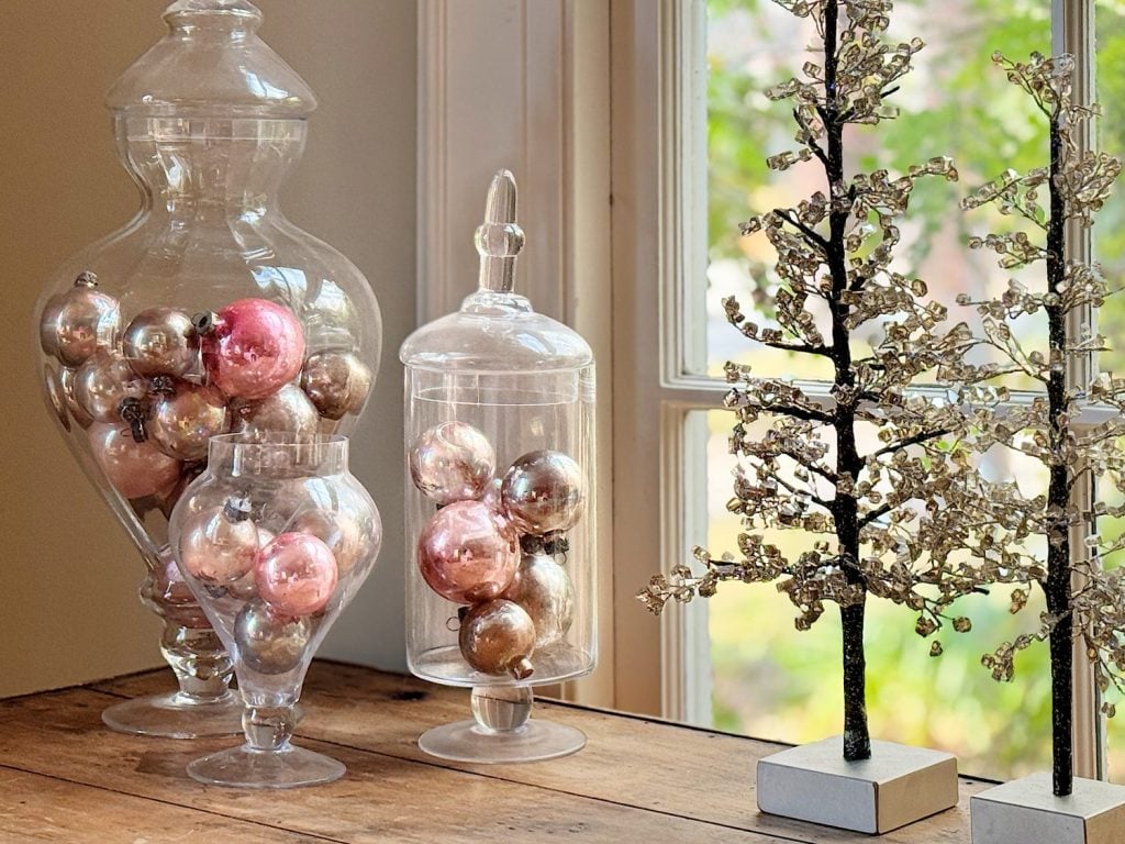 Glass jars filled with pink and silver ornaments are placed on a wooden table beside a decorative tree by a window.