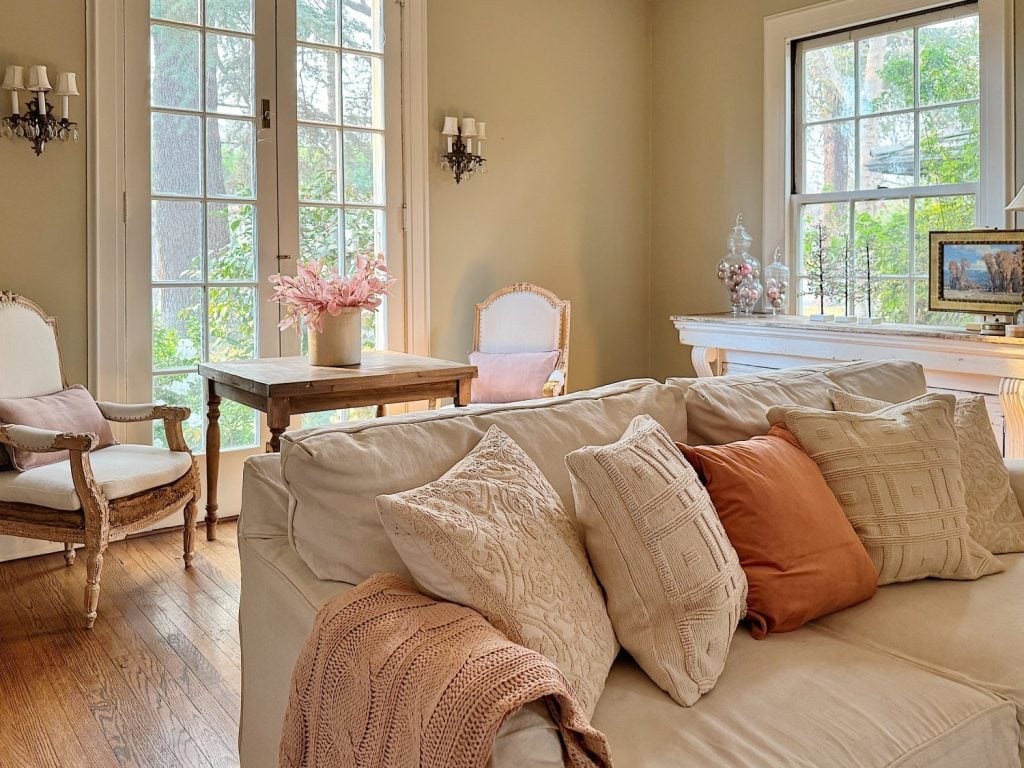 Living room with a beige sofa, assorted pillows, a wooden floor, and a small table with pink flowers. Large windows and light-colored walls create a bright, airy atmosphere.