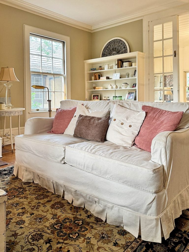 Cozy living room with a white couch, pink and beige pillows, a rug, and a bookshelf filled with decor and photos. A window and side table with a lamp are in the background.