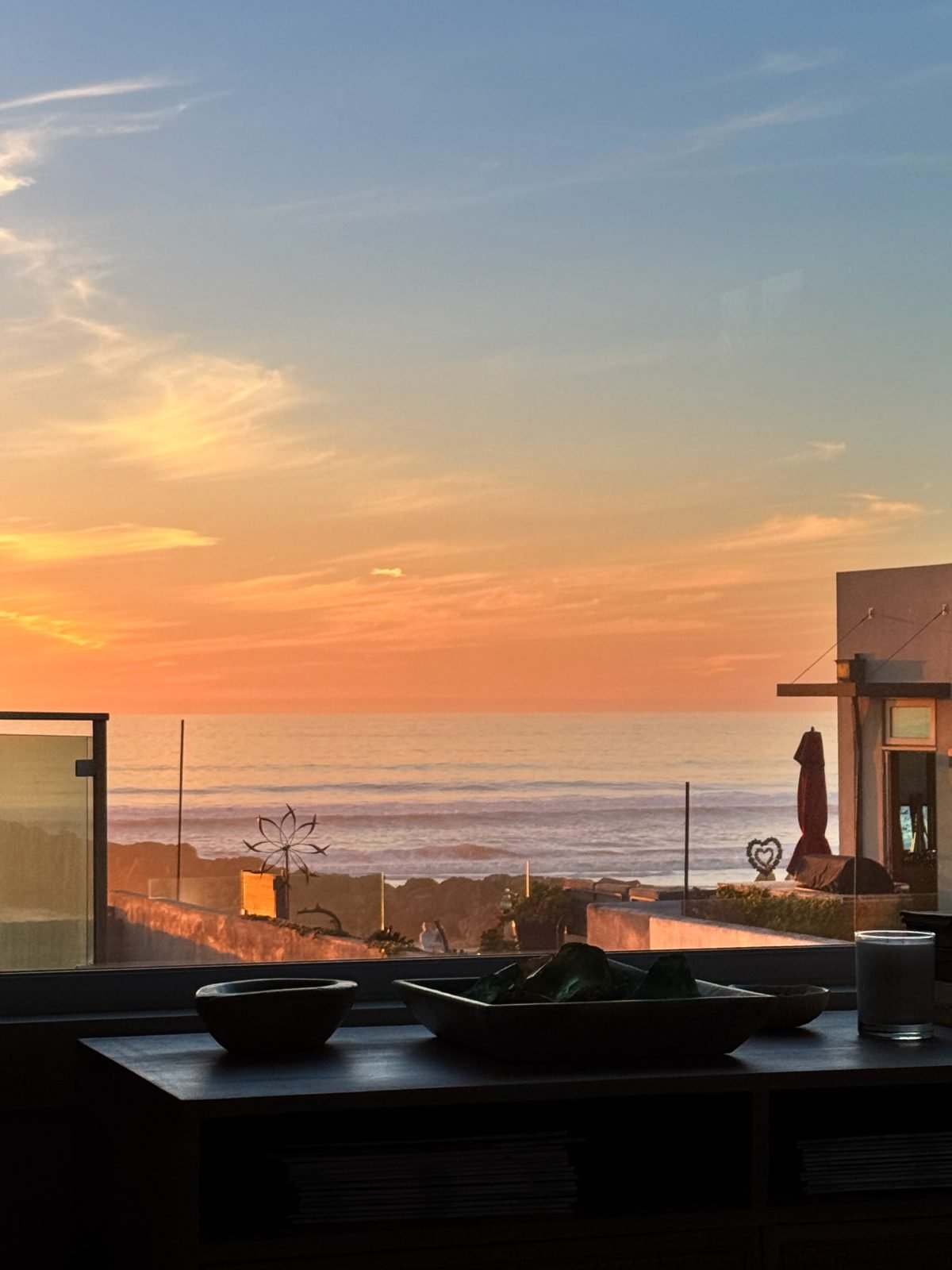 Sunset view from a window with a beach and ocean in the background. Silhouetted objects, including a patio umbrella and decor, are in the foreground.