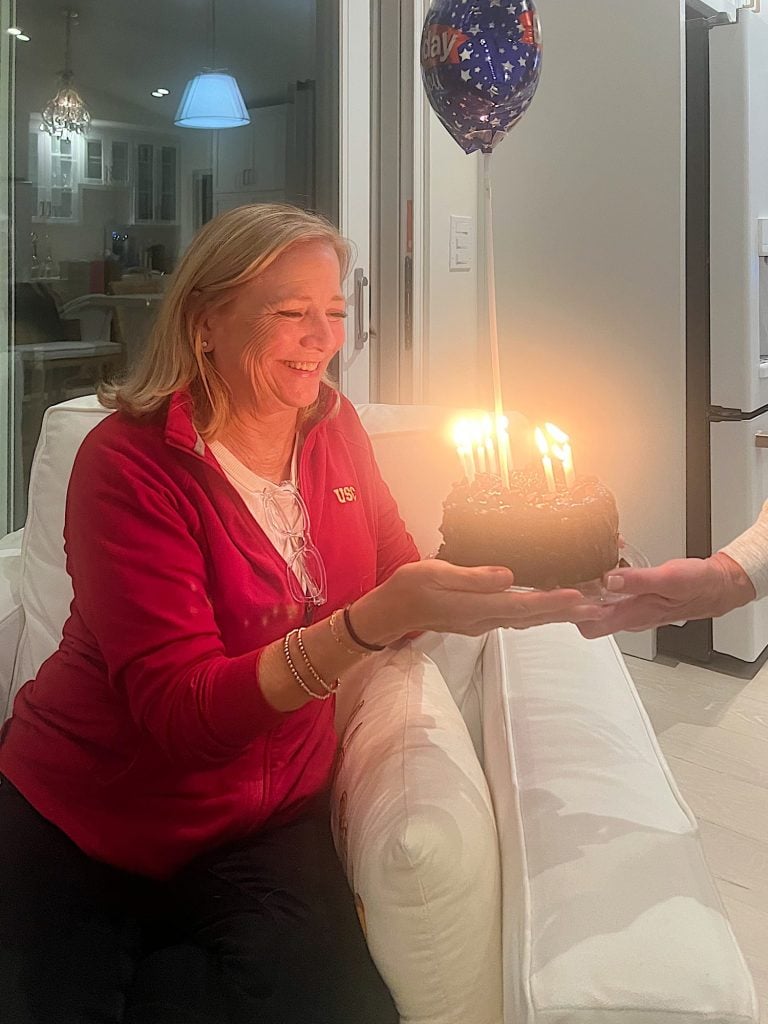 A smiling woman in a red jacket holds a small chocolate cake with lit candles and a star balloon inside a cozy room.