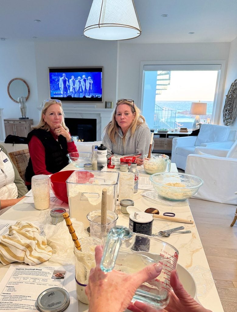 People sitting at a table covered with baking ingredients, utensils, and a recipe in a bright kitchen with a view of the ocean outside. A TV is on in the background.