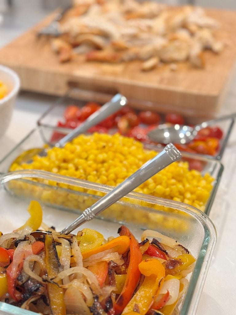 A variety of foods including sautéed peppers and onions, corn, and halved cherry tomatoes in dishes, with sliced chicken on a cutting board in the background.