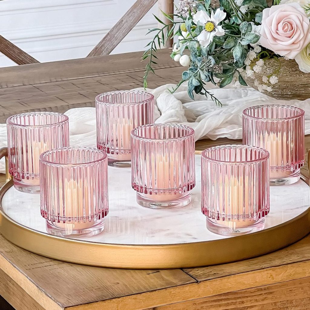 Six pink ribbed glass candle holders with lit candles on a round golden tray, placed on a wooden table with floral decorations in the background.