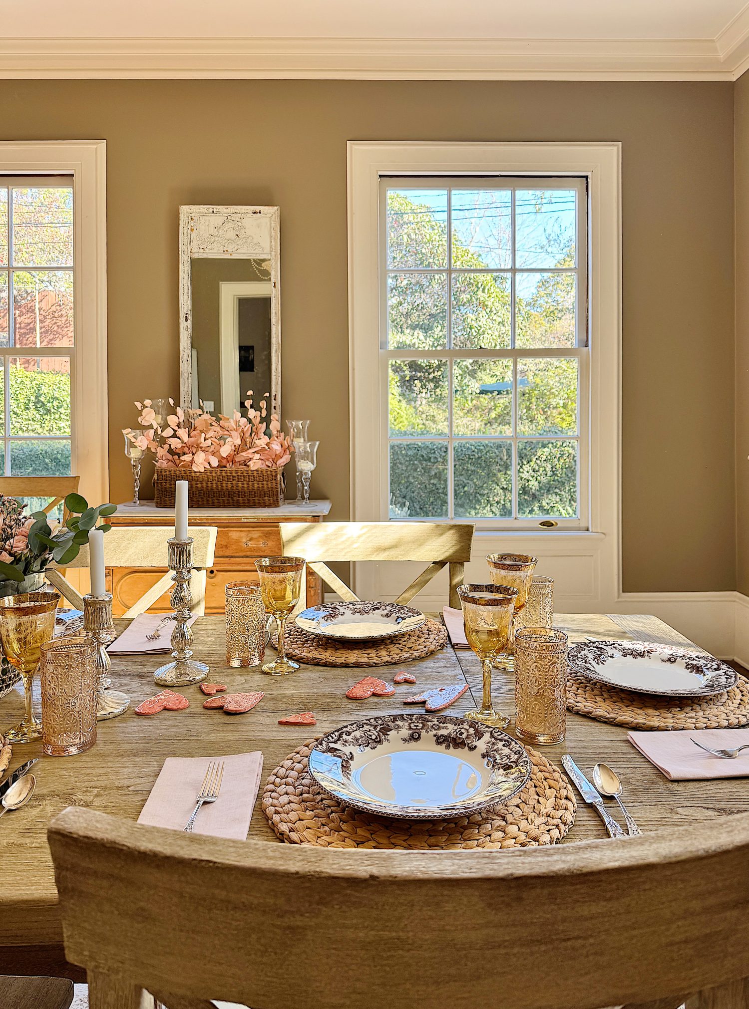 A dining table set for a meal with woven placemats, plates, glasses, cutlery, candles, and flower decorations in a room with large windows and beige walls.