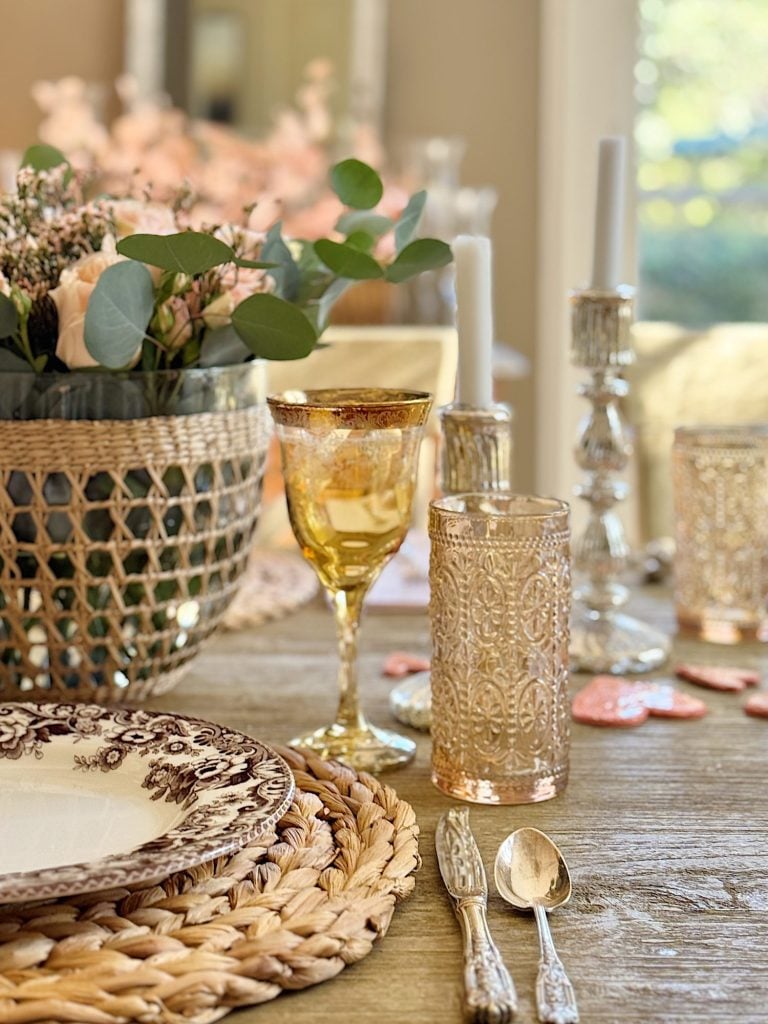 A table set with elegant glassware, floral centerpiece, and intricately patterned plates on woven placemats. Two lit candles are in the background.