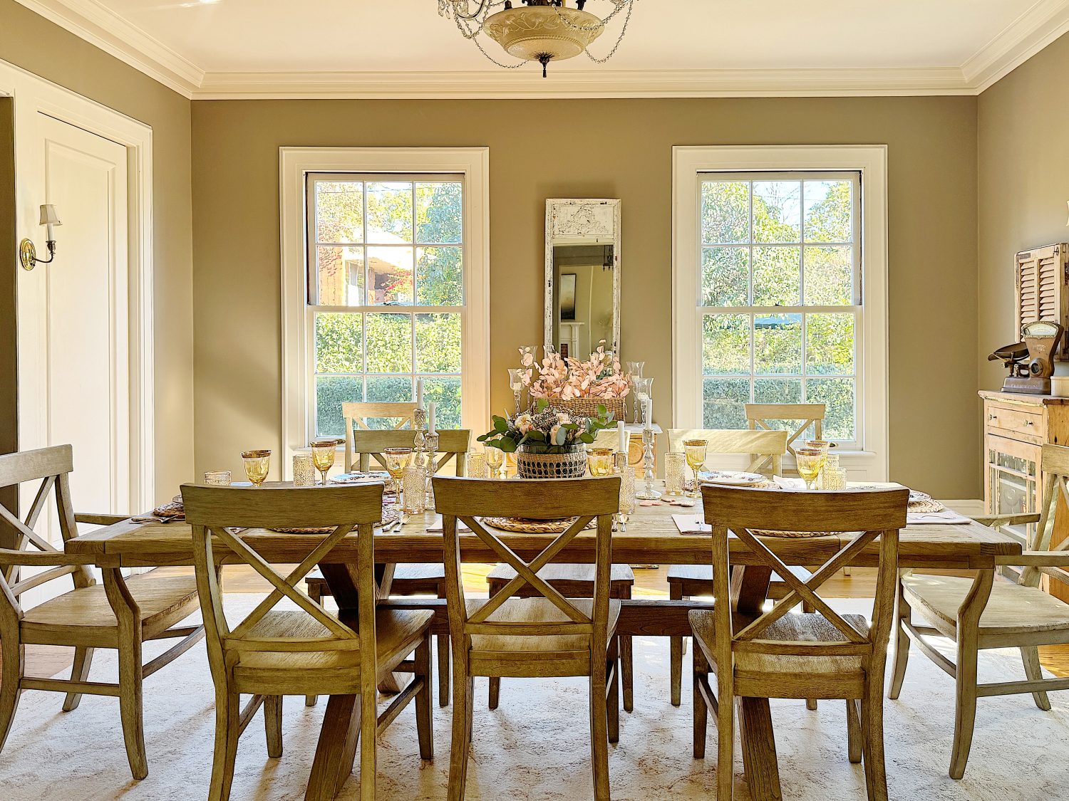 A sunlit dining room with a wooden table set for eight, featuring floral centerpieces and glassware. Three large windows brighten the beige-colored walls.