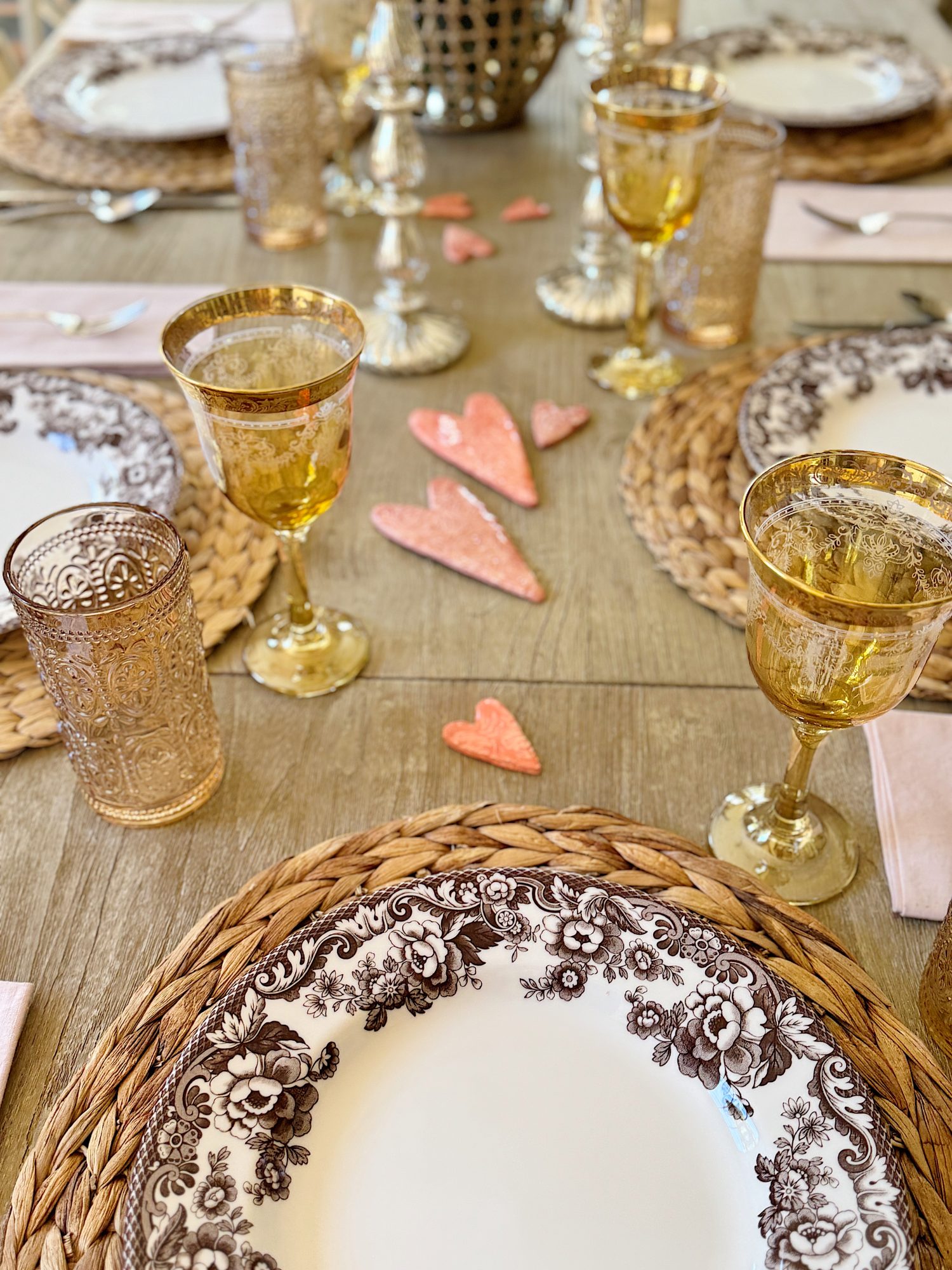 A dining table is set with floral-patterned plates, gold-rimmed glasses, beige woven placemats, pink napkins, and decorative pink heart ornaments.