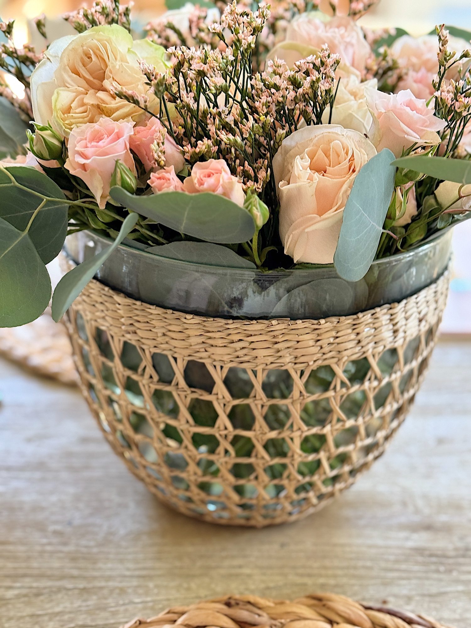 A woven basket vase holds a floral arrangement of pink and cream roses with green leaves and small pink buds, placed on a wooden table.