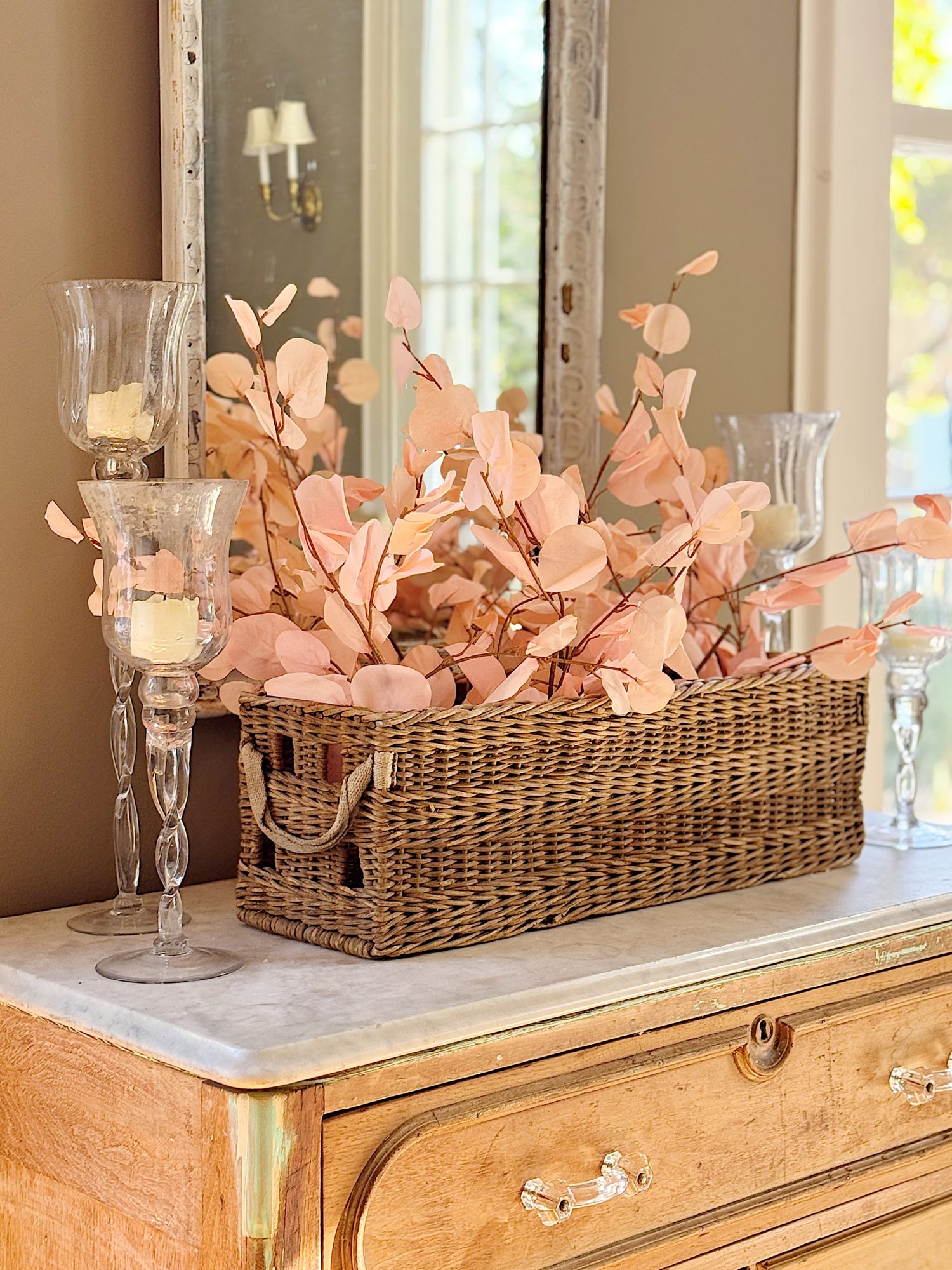 Wicker basket with pink foliage on a wooden dresser, accompanied by glass candle holders and a mirror.