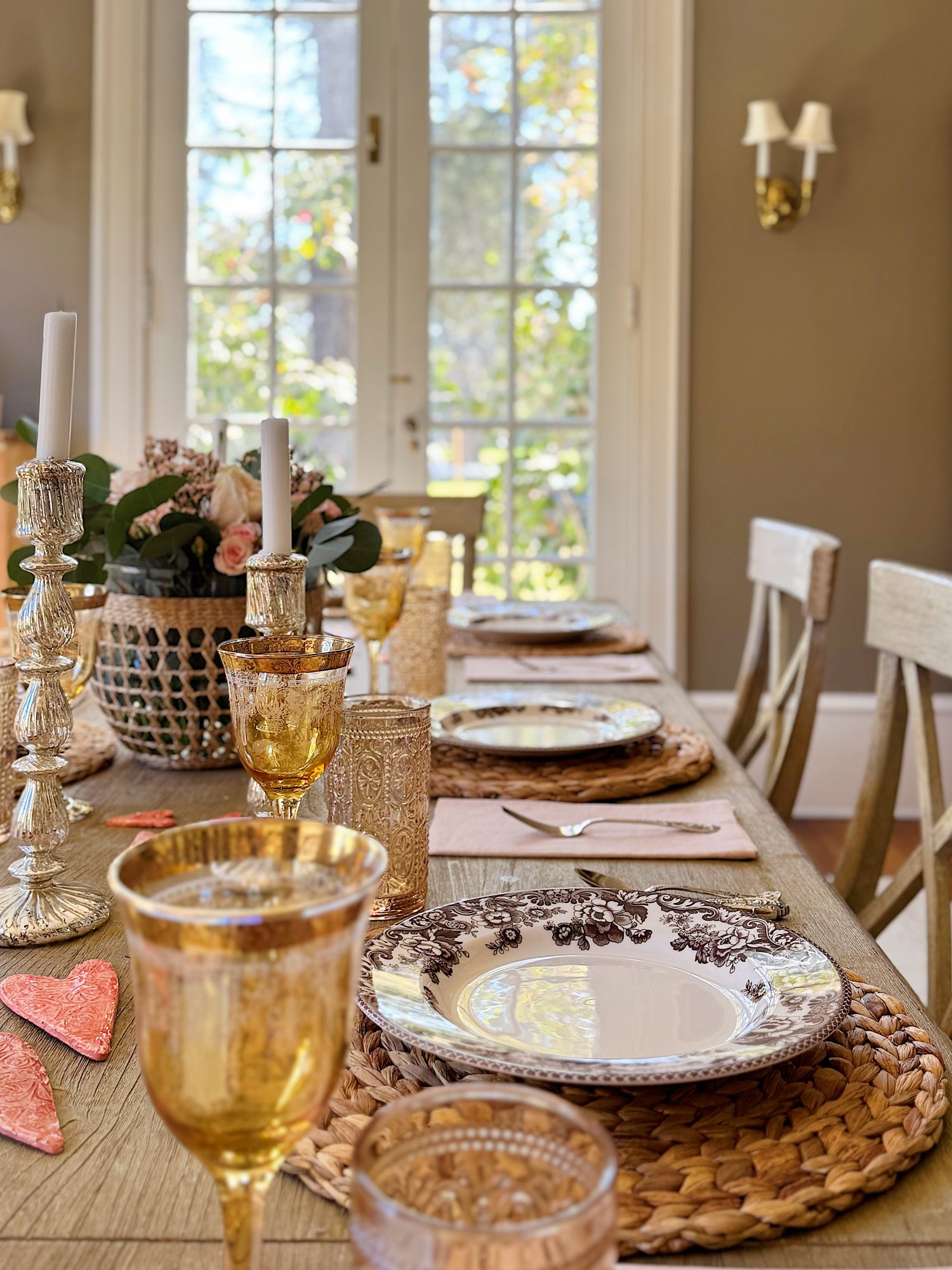 A dining table set with decorative plates, woven placemats, amber glasses, and lit candles. A bouquet of flowers is in the background.
