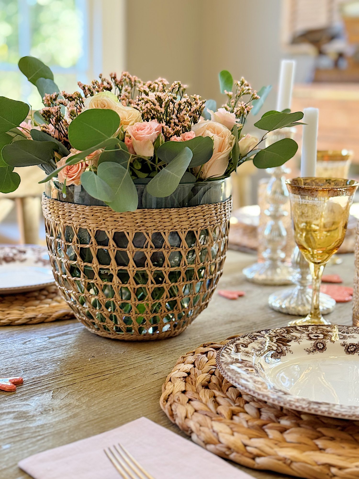 A woven basket with flowers on a wooden dining table, surrounded by dinner plates, woven placemats, and crystal glasses.