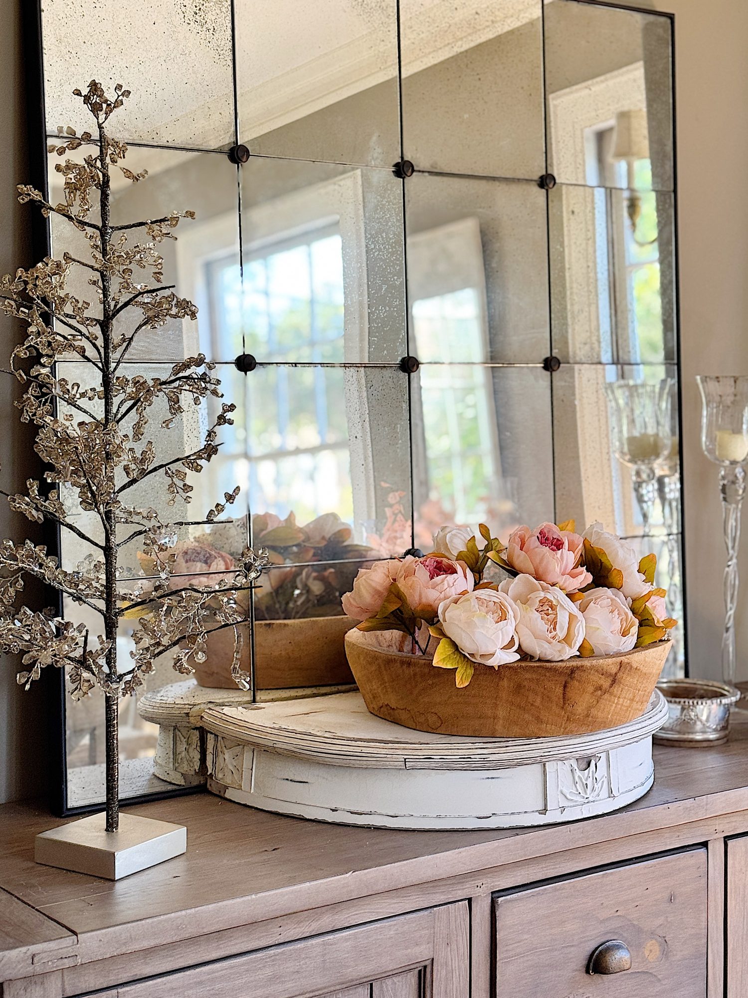 A wooden bowl with pink flowers sits on a white mantelpiece in front of a large mirror. A decorative tree and candle holder are on either side.