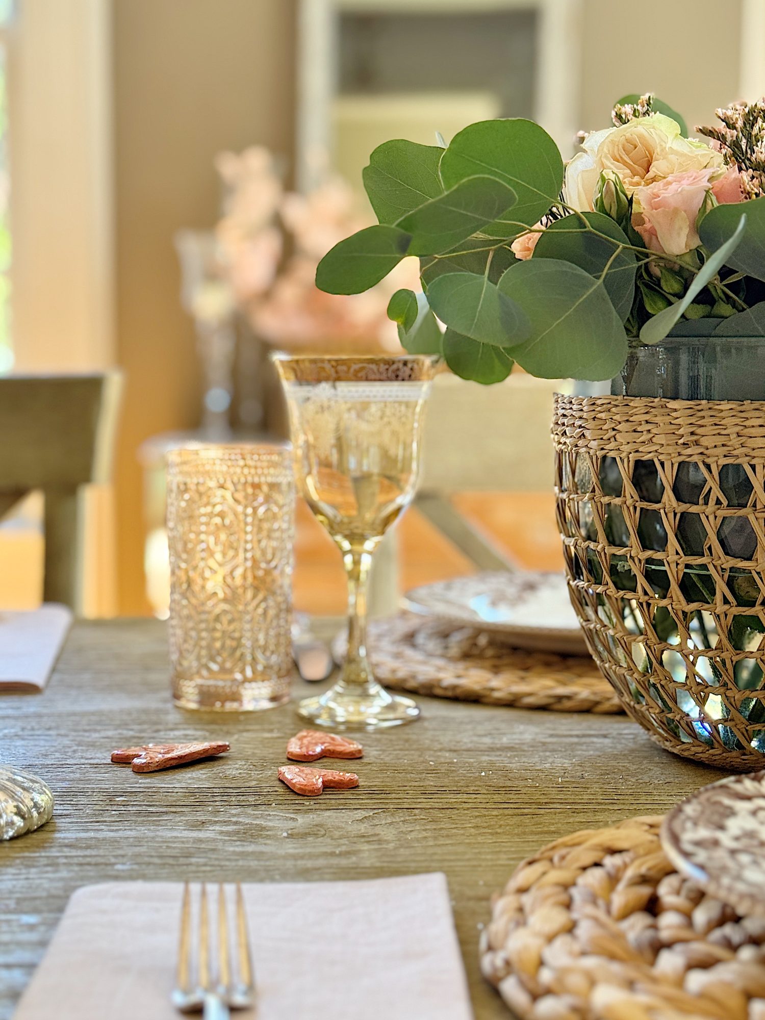 A dining table set with woven placemats, glassware, and cutlery, featuring a large vase with green leaves and flowers. Small decorative items are placed beside the glass.