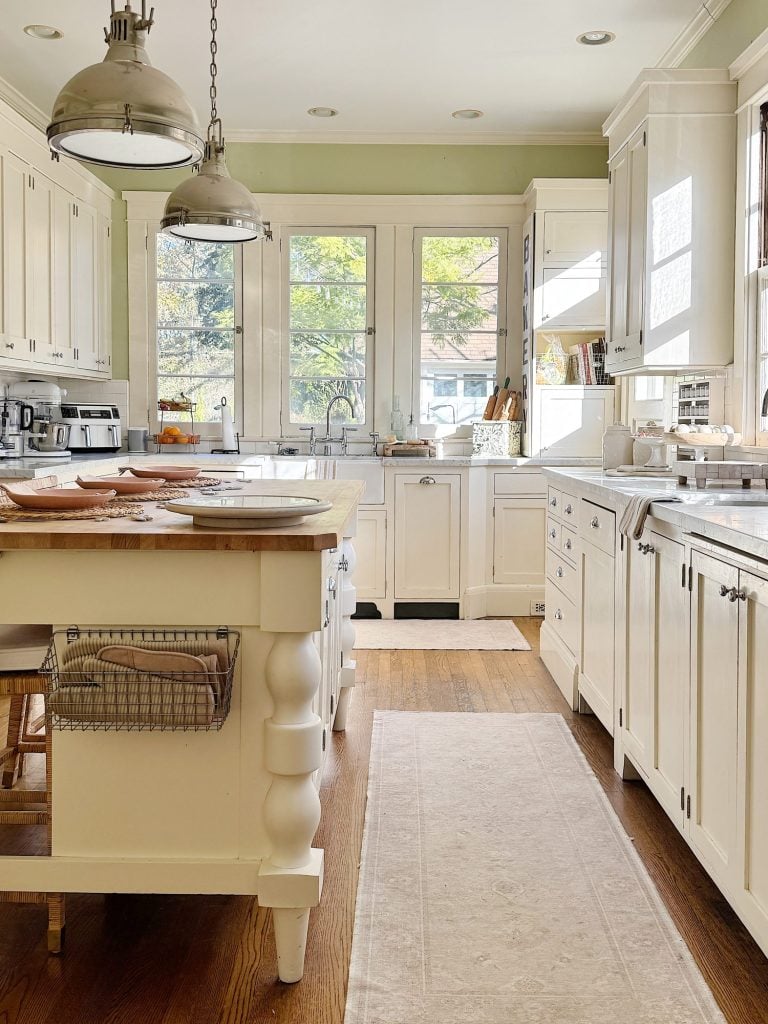 A bright kitchen with white cabinets, wooden countertops, and stainless steel appliances. Two pendant lights hang above a central island with stools. Natural light filters through large windows.