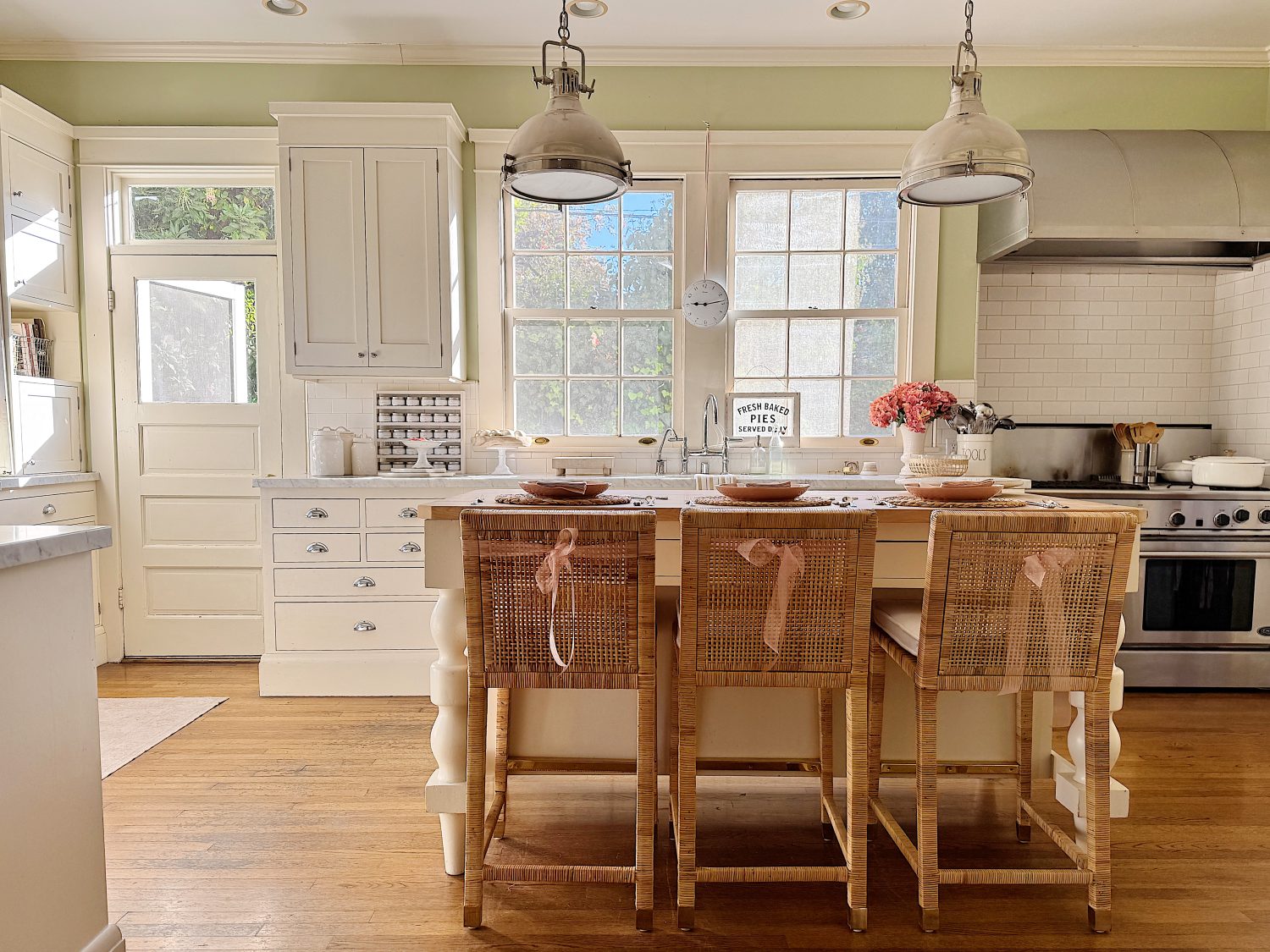 A bright kitchen with a wooden island, wicker chairs, white cabinets, and stainless steel appliances. Pink flowers and a wall clock are visible, with windows letting in natural light.