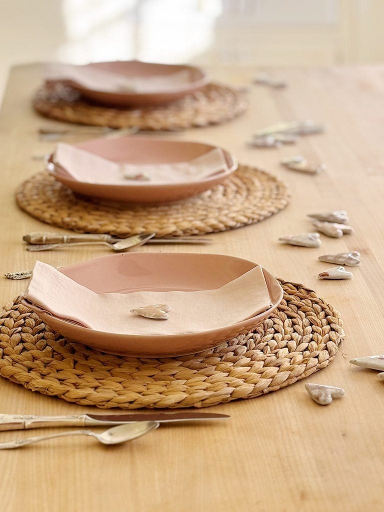 A wooden dining table set with three woven placemats, pink ceramic plates, and cloth napkins. Small heart-shaped stones are scattered around the place settings.