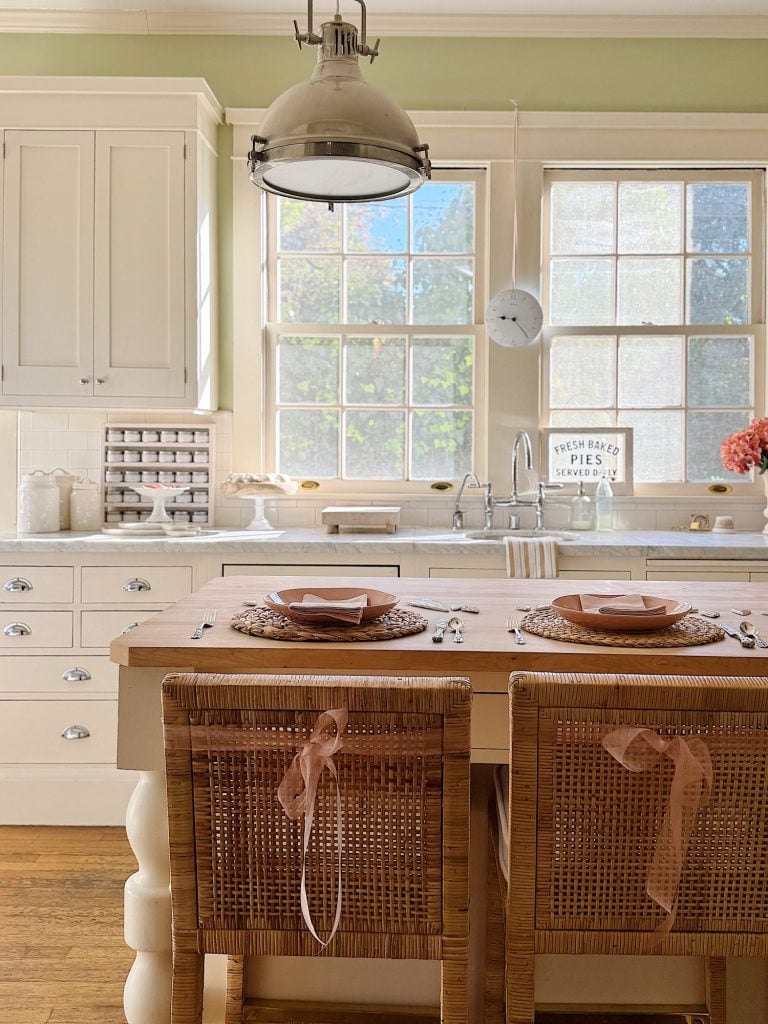 A bright kitchen with a wooden island, wicker chairs, pendant light, and white cabinets. Two place settings on the island feature woven placemats and neutral dishware.