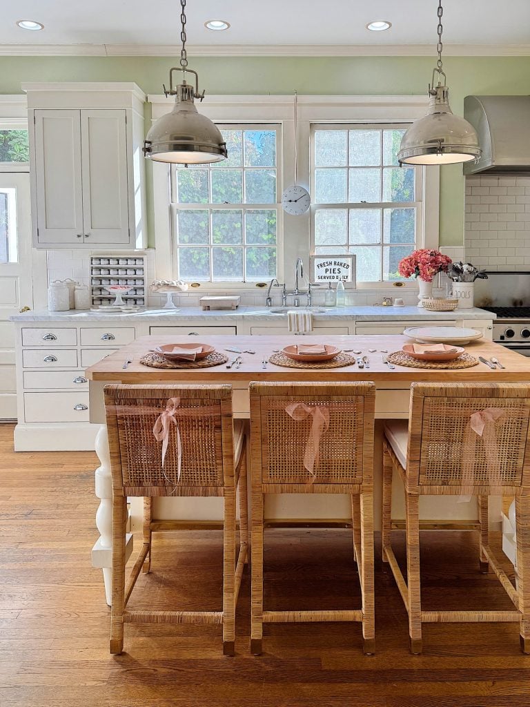 A bright kitchen with a wooden island, wicker bar stools with pink bows, white cabinetry, and overhead pendant lights. Pink flowers and tableware adorn the counter.