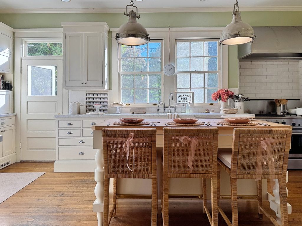 A kitchen with a central island and three chairs, pendant lights, a stove, cabinets, and windows. Pink ribbons adorn the chair backs, and pink flowers decorate the table.