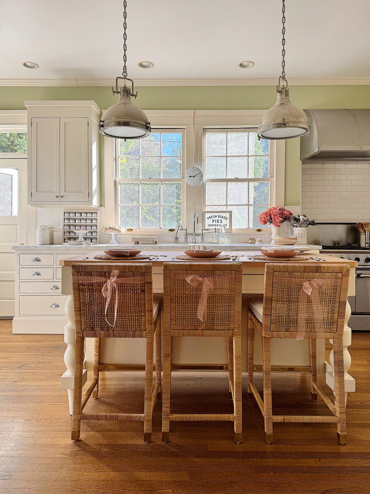 A kitchen with a central island featuring three wicker chairs, pendant lights, and a vase of flowers. White cabinets, a farmhouse sink, and a stainless steel range are visible.