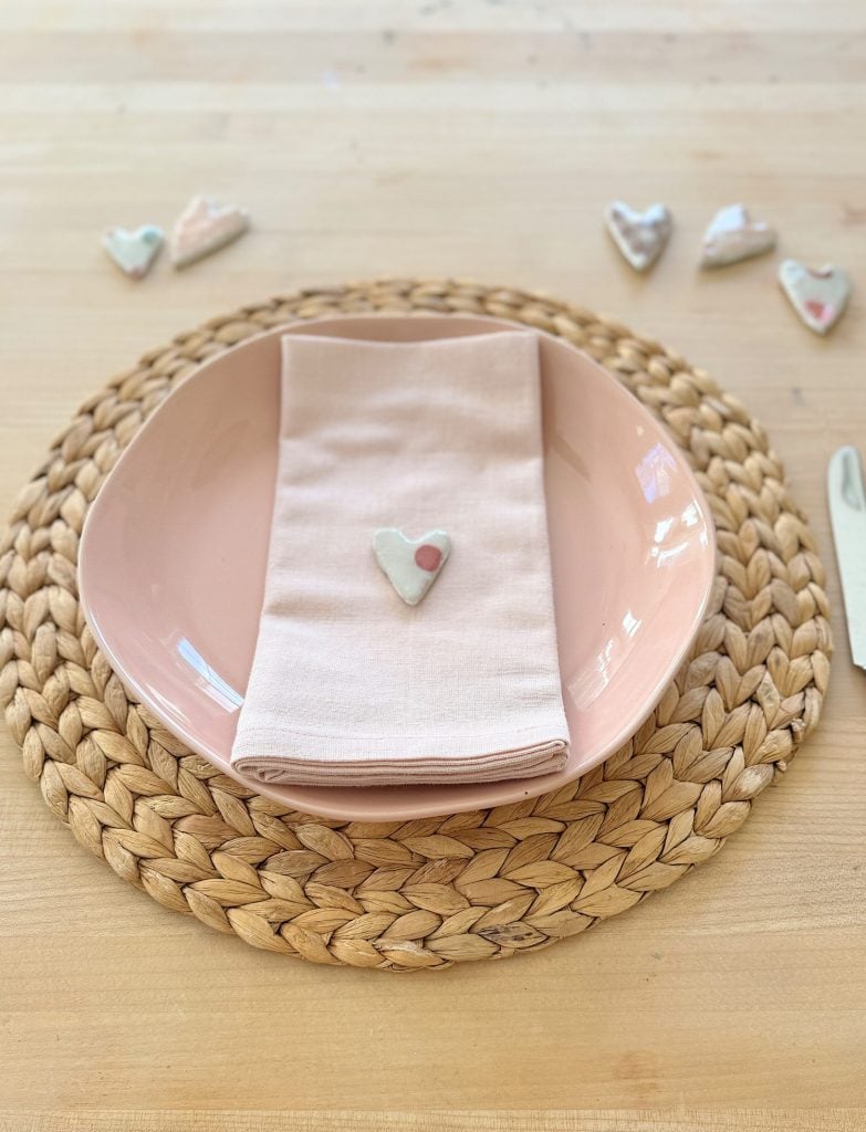 Pink plate on a woven placemat with a folded napkin topped by a heart-shaped ornament. Heart decorations are scattered in the background.