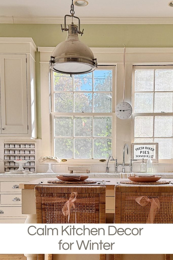 Bright kitchen with large windows, wicker bar stools, and a metal pendant light. Shelves and countertop accessories add a cozy touch. Text overlay reads: "Calm Kitchen Decor for Winter.