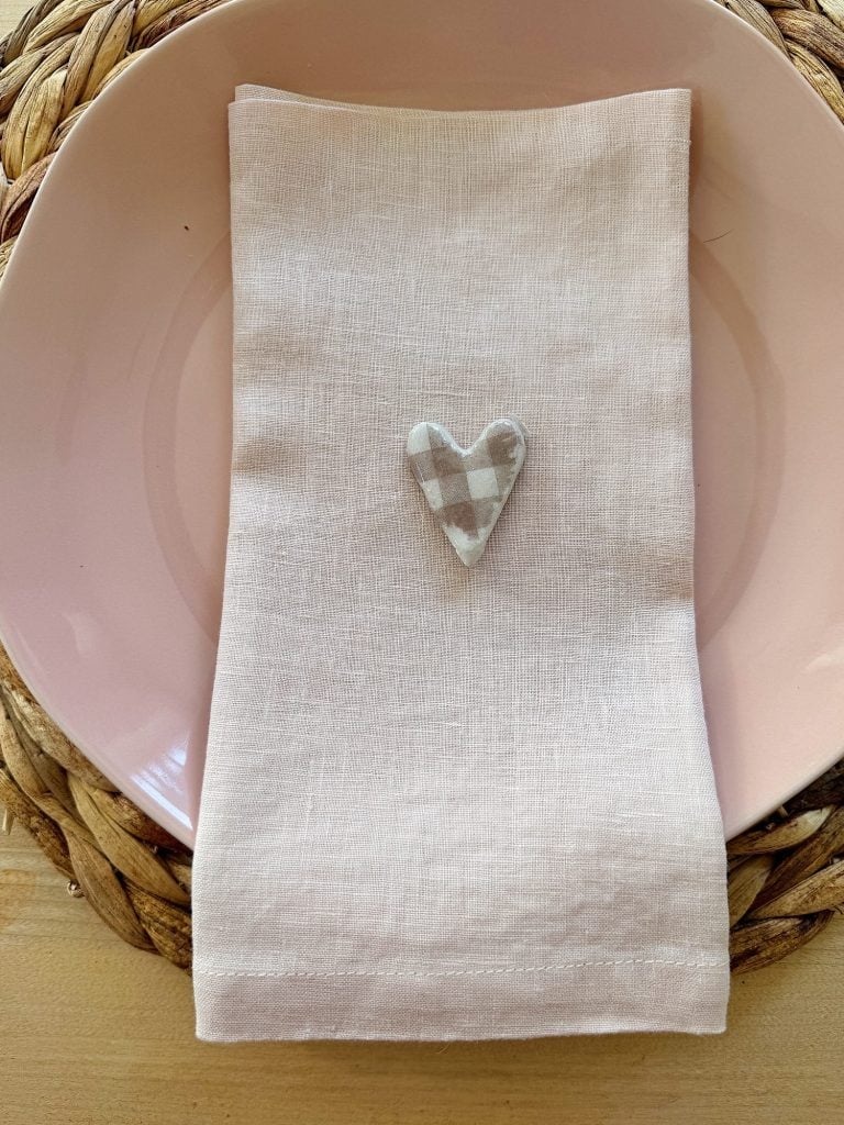 Pink plate with a folded beige napkin topped with a small checkered heart decoration, placed on a woven placemat.