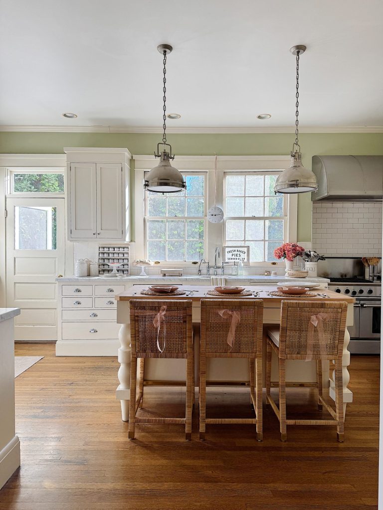 A bright kitchen with wooden flooring, an island with wicker chairs, pendant lights, and white cabinets. A door and large windows let in natural light. Pink flowers adorn the counter.