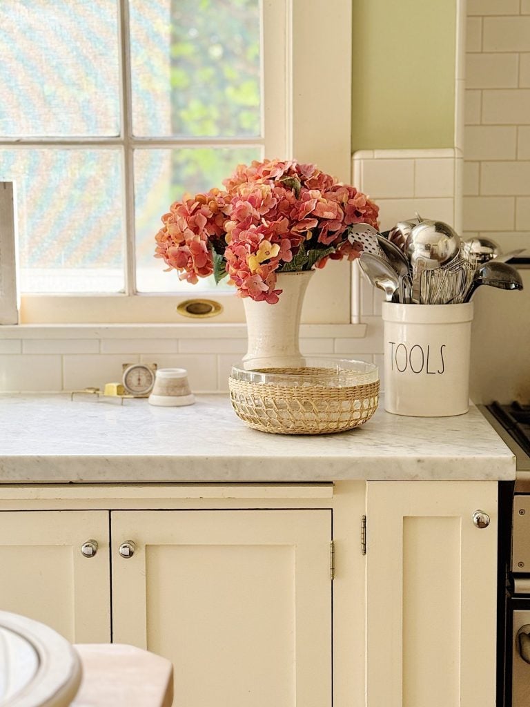 Kitchen counter with a vase of pink flowers, a golden bowl, and a container labeled "TOOLS" holding kitchen utensils, near a window.