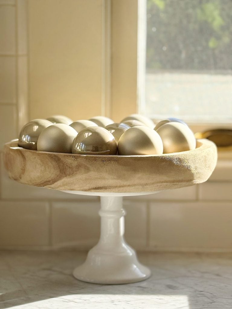 A wooden bowl on a white pedestal holds shiny golden spheres, placed on a marble countertop with sunlight streaming in from a window.