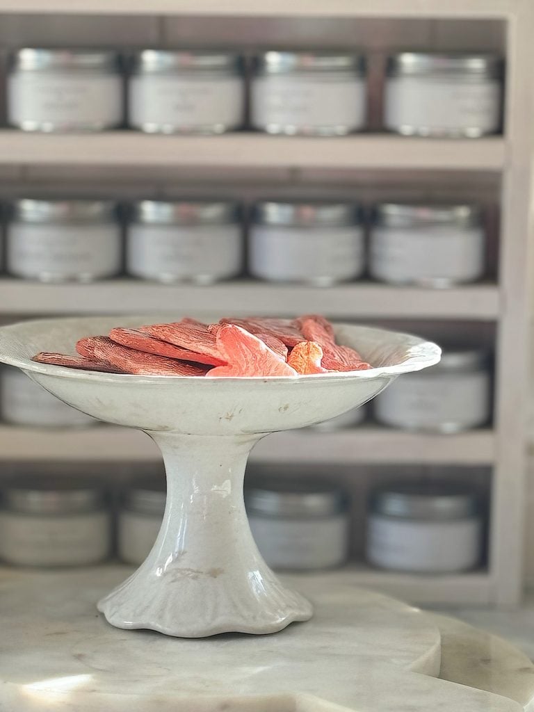 A ceramic pedestal bowl with slices of dried fruit in front of shelves filled with jars.