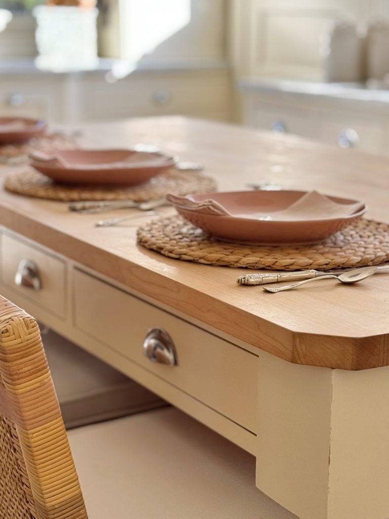 A wooden dining table with three place settings, featuring woven placemats, brown bowls, and silverware, in a well-lit kitchen.