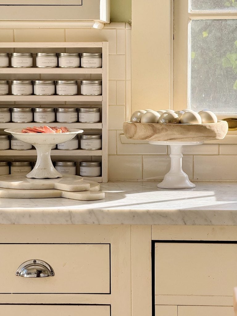 A kitchen counter with spice jars, a cake stand holding eggs, another holding food, and a sunlit window.