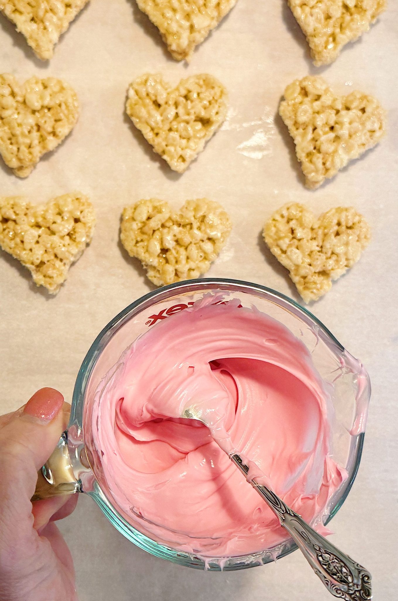 A hand holds a glass bowl of pink frosting with a spoon. Heart-shaped rice cereal treats are arranged on parchment paper in the background.