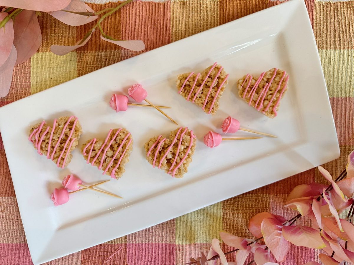 Heart-shaped treats with pink drizzle on a rectangular white plate, accompanied by toothpick rose decorations. Set on a checked tablecloth with pink leaves.