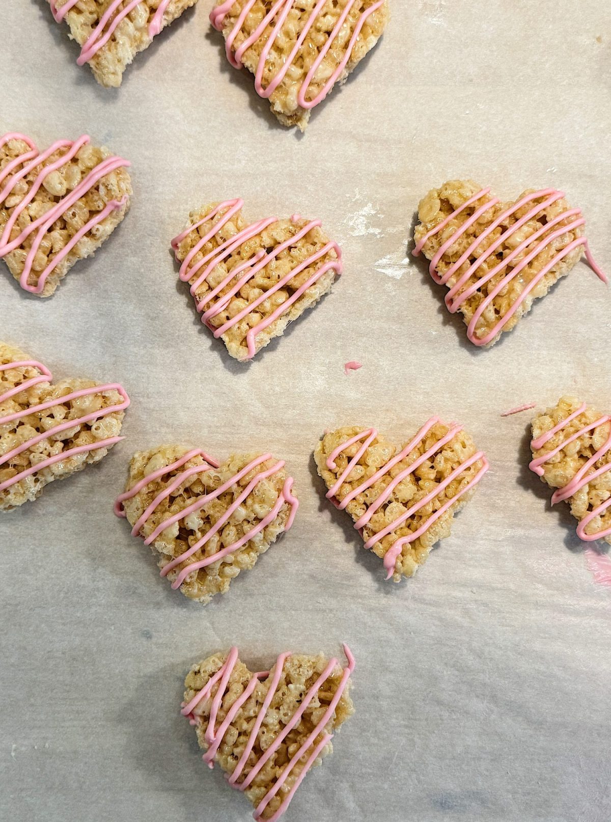 Heart-shaped rice crispy treats drizzled with pink icing, arranged on parchment paper.