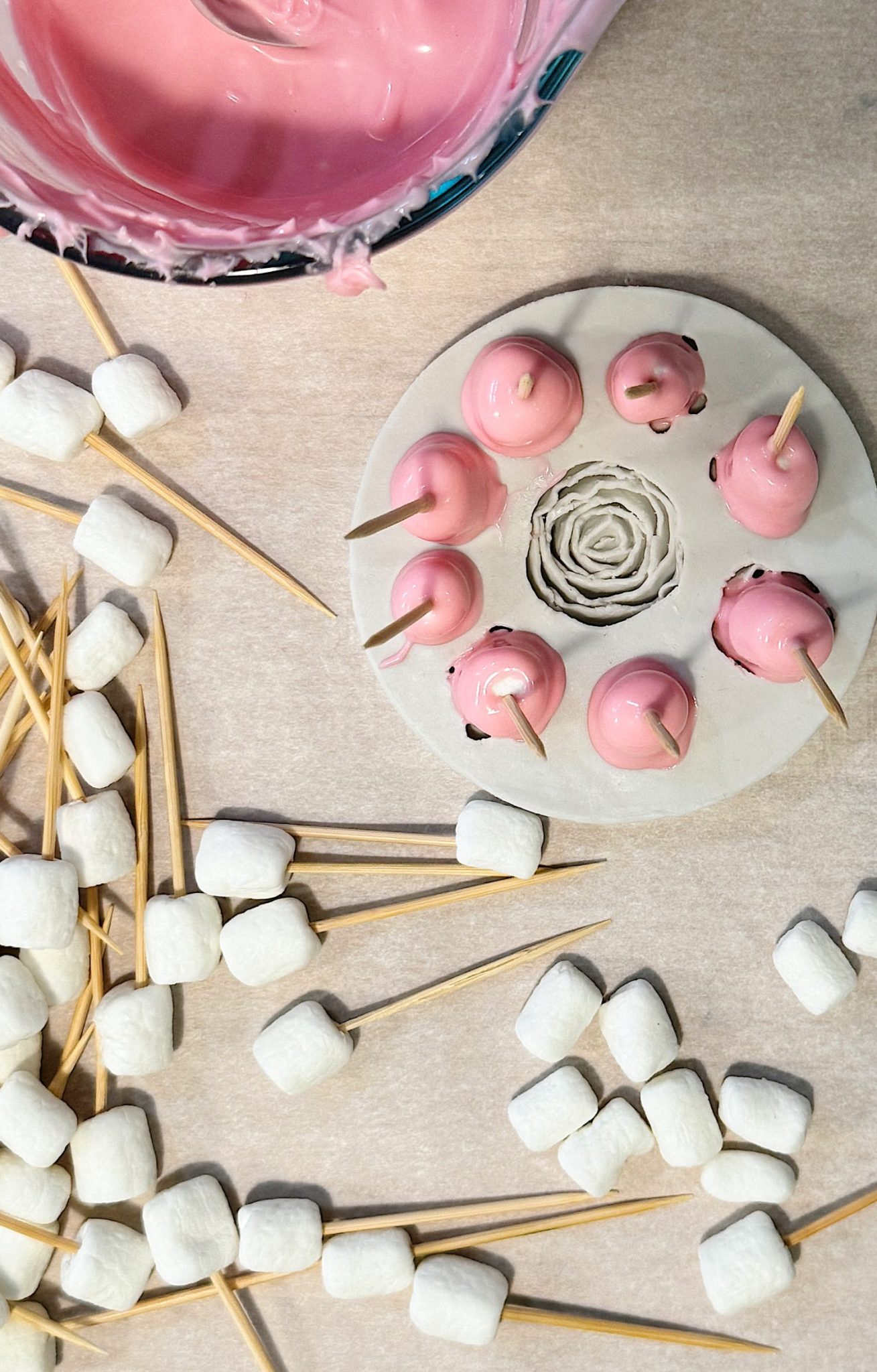 Pink-coated cake pops drying on a stand, surrounded by marshmallows on toothpicks on a parchment-lined surface.