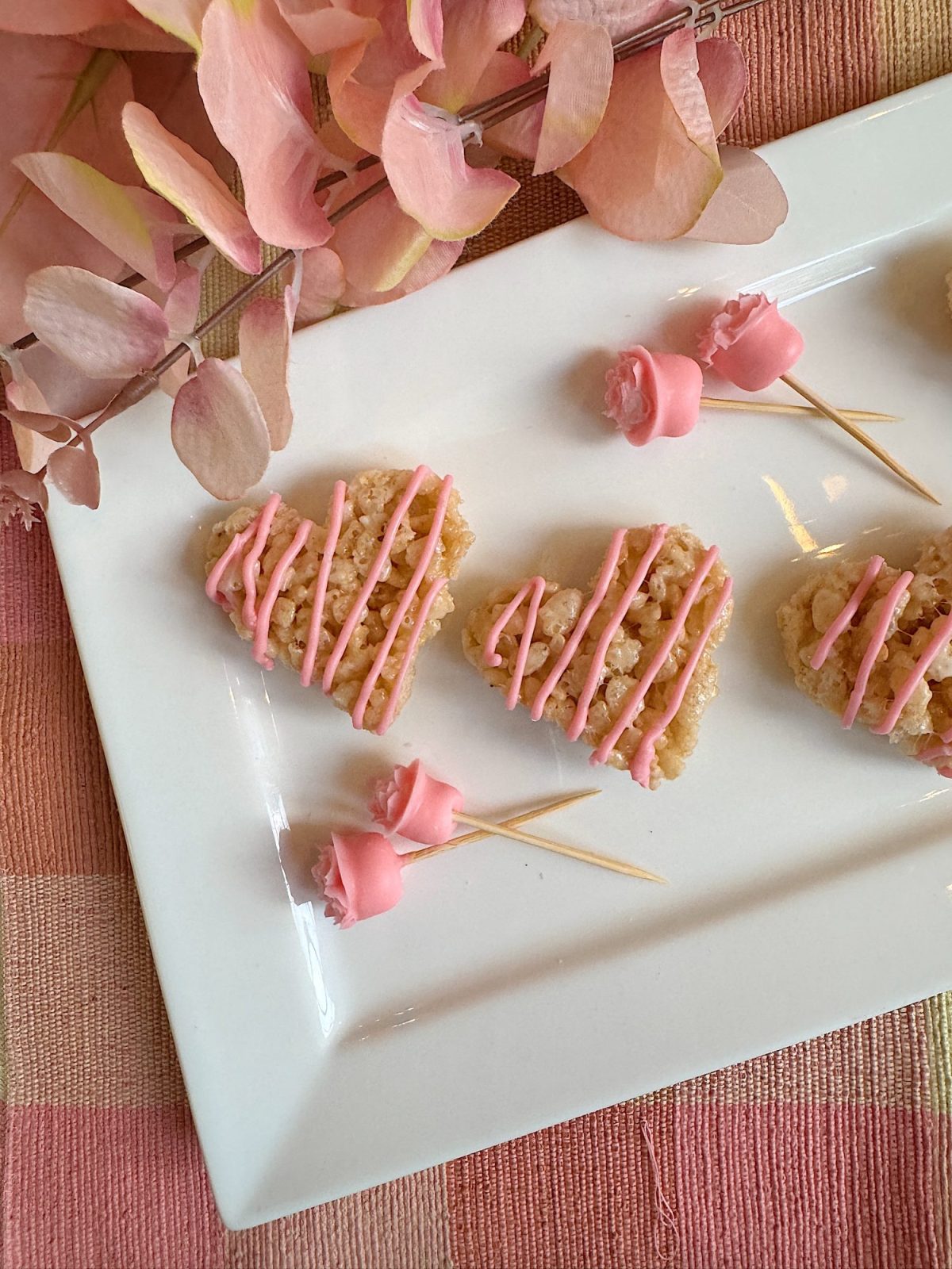 Heart-shaped rice treats with pink drizzle on a white plate, accompanied by pink rose-shaped toothpicks, arranged on a pink-patterned tablecloth.
