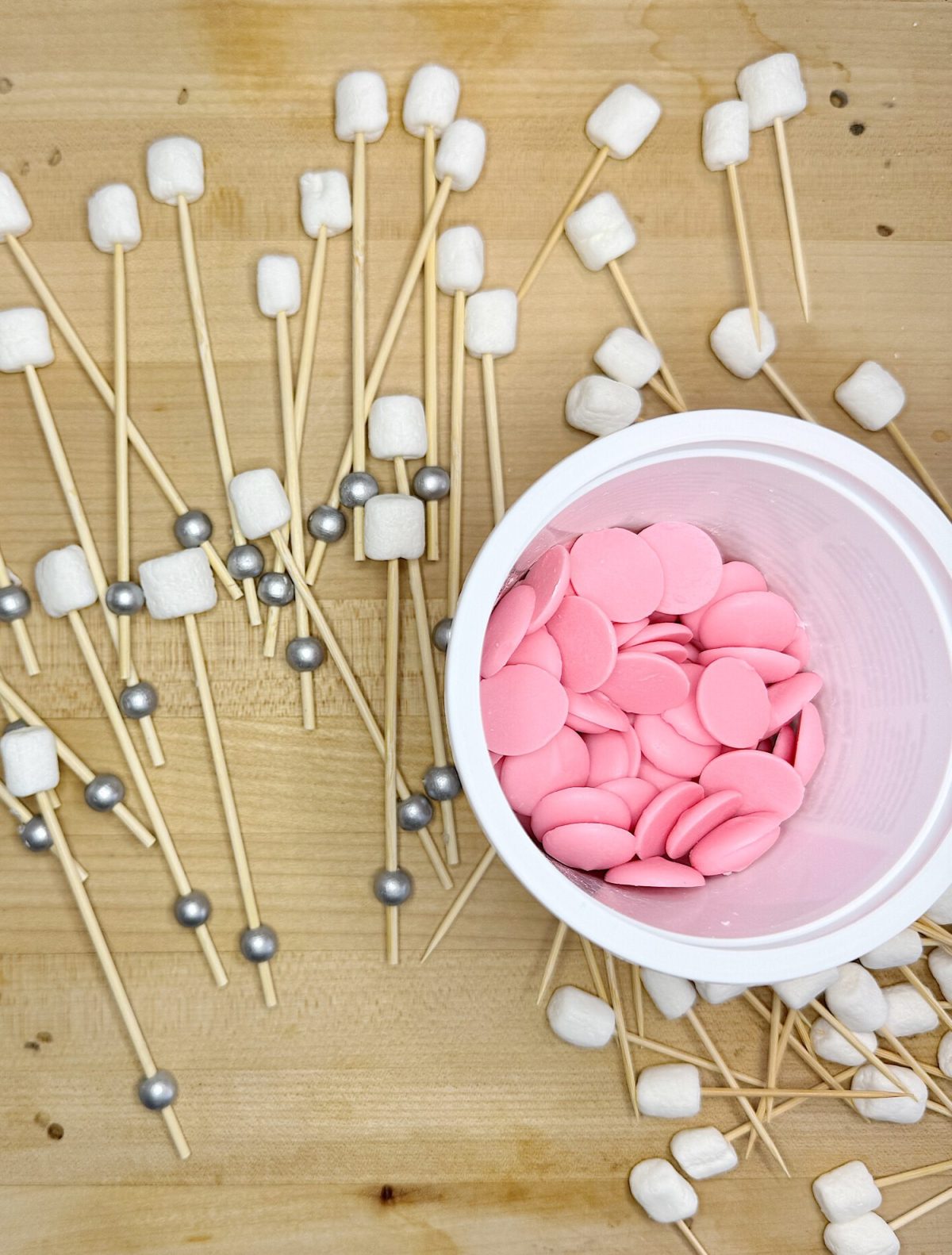 A white container with pink candy melts, surrounded by marshmallows and silver candies on skewers, placed on a wooden surface.