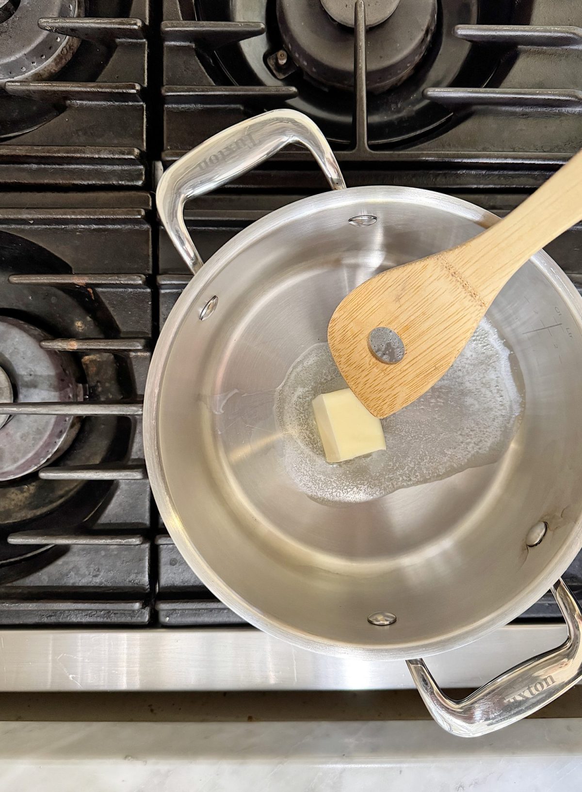 A wooden spoon stirring a melting butter cube in a stainless steel saucepan on a stovetop.