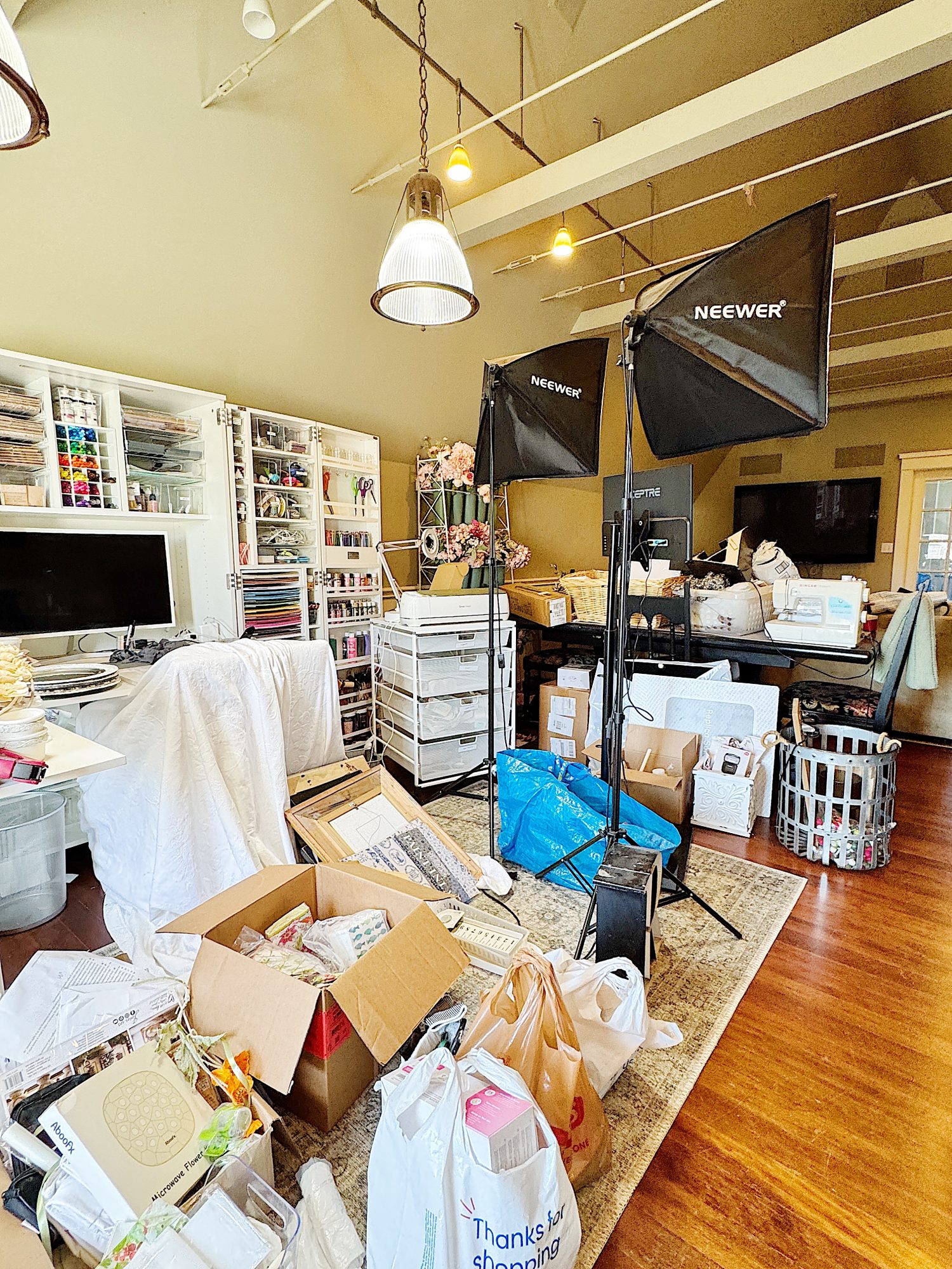 A cluttered room with open boxes, bags, and art supplies. Softbox lights are set up, and shelves hold various materials. A computer is on the desk.