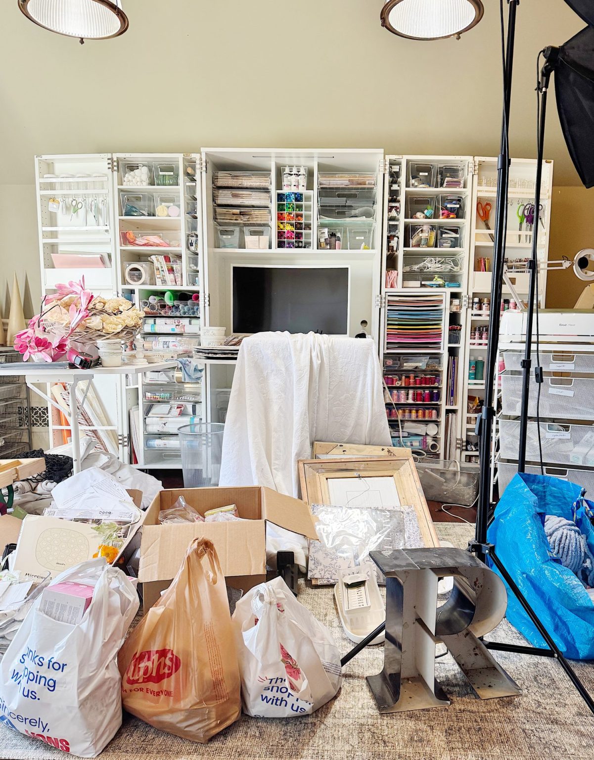 Cluttered craft room with shelves full of supplies, bags on the floor, a large white shelf unit, a TV, and scattered items including cardboard boxes and a metal letter 'P'.