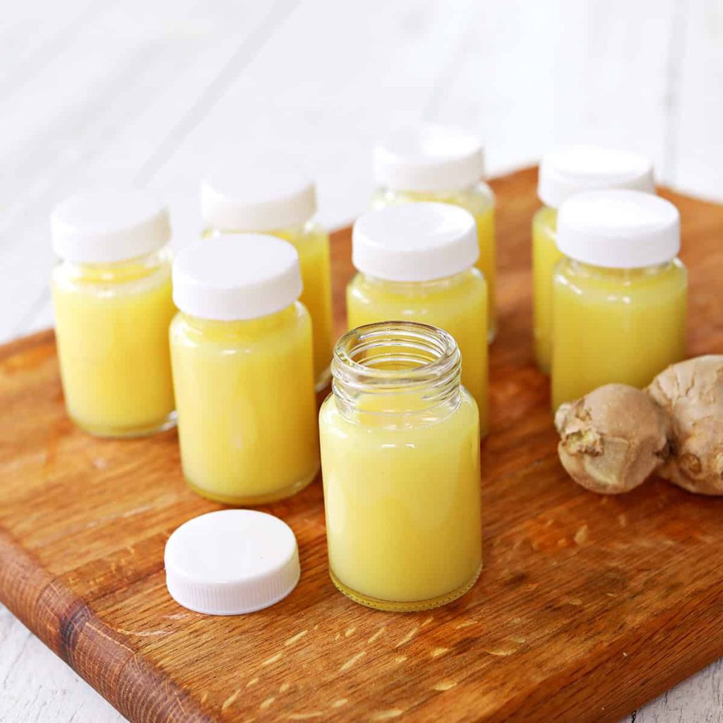 Eight small jars of yellow liquid with white lids on a wooden board, next to pieces of ginger.