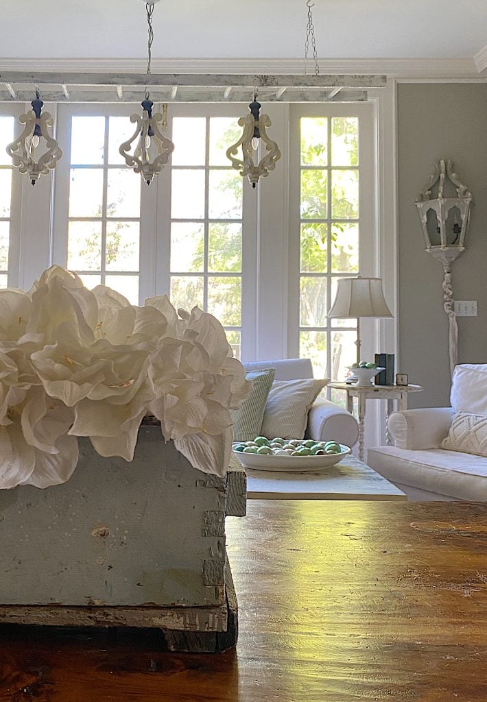 A cozy living room with large windows and a winter color palette features white sofas, a wooden table adorned with white flowers, and a lamp. Decorative light fixtures hang from the ceiling, while a bowl of green objects adds contrast on the side table.