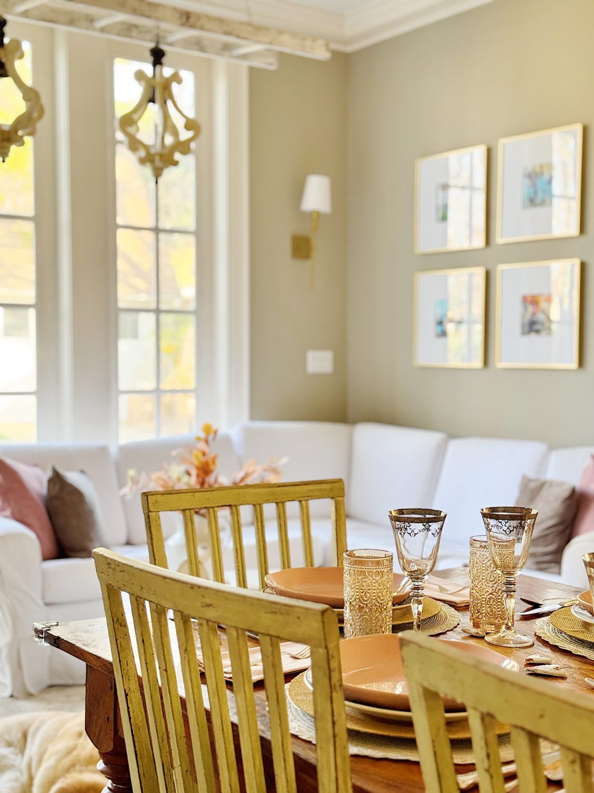 A cozy living room with a white sectional sofa. A dining table in the foreground is set with orange plates and glassware. Four framed pictures hang on the wall, and windows let in natural light.