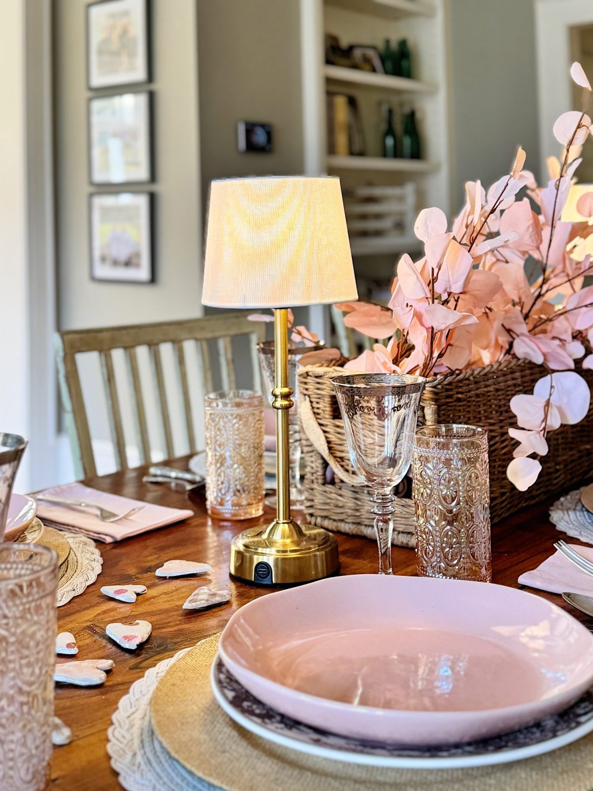 Elegant dining table setup with a small lamp, pink plates, decorative glasses, and pink floral arrangement in a basket.