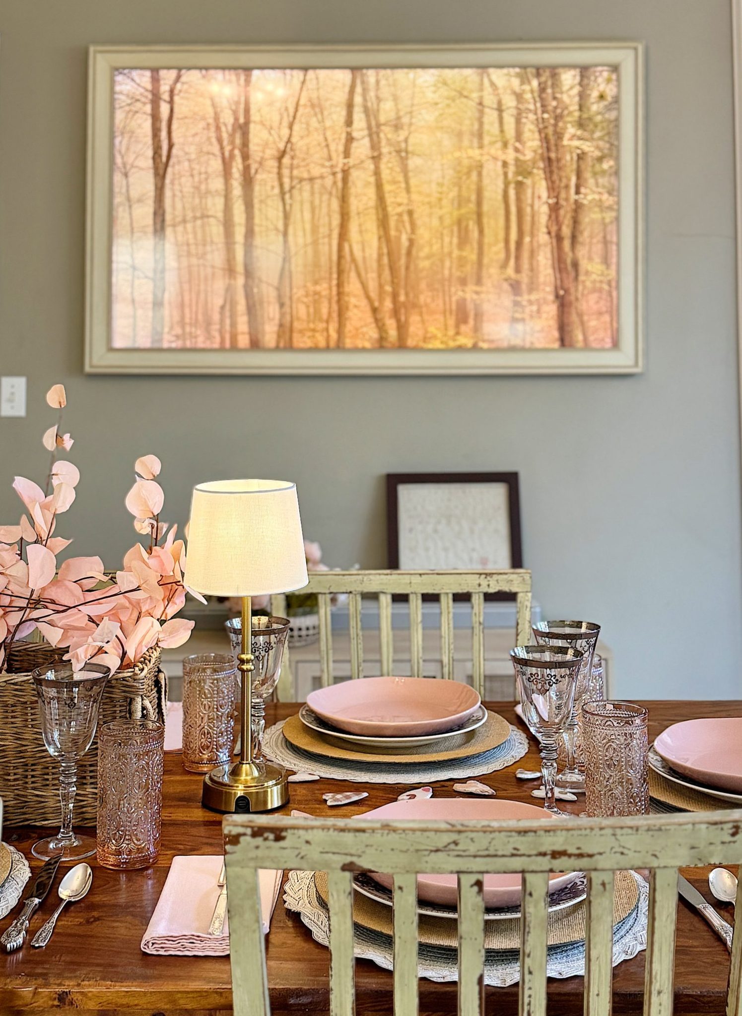A dining table set with pink plates, glassware, and a small lamp. Pink flowers and a woven basket are on the table. A framed forest picture hangs on the wall.